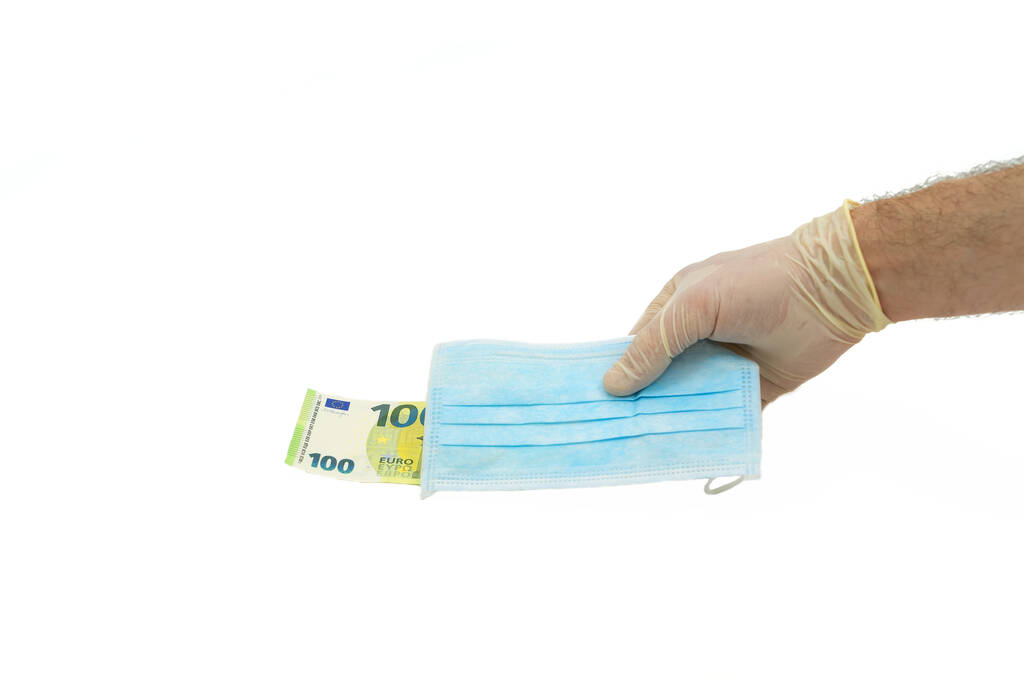 Une main dans un gant de protection blanc donne un masque médical bleu pour de l'argent de 100 euros. Concept d'achat d'un masque en Europe, protection contre le virus. Fond blanc isolé
. - Photo, image