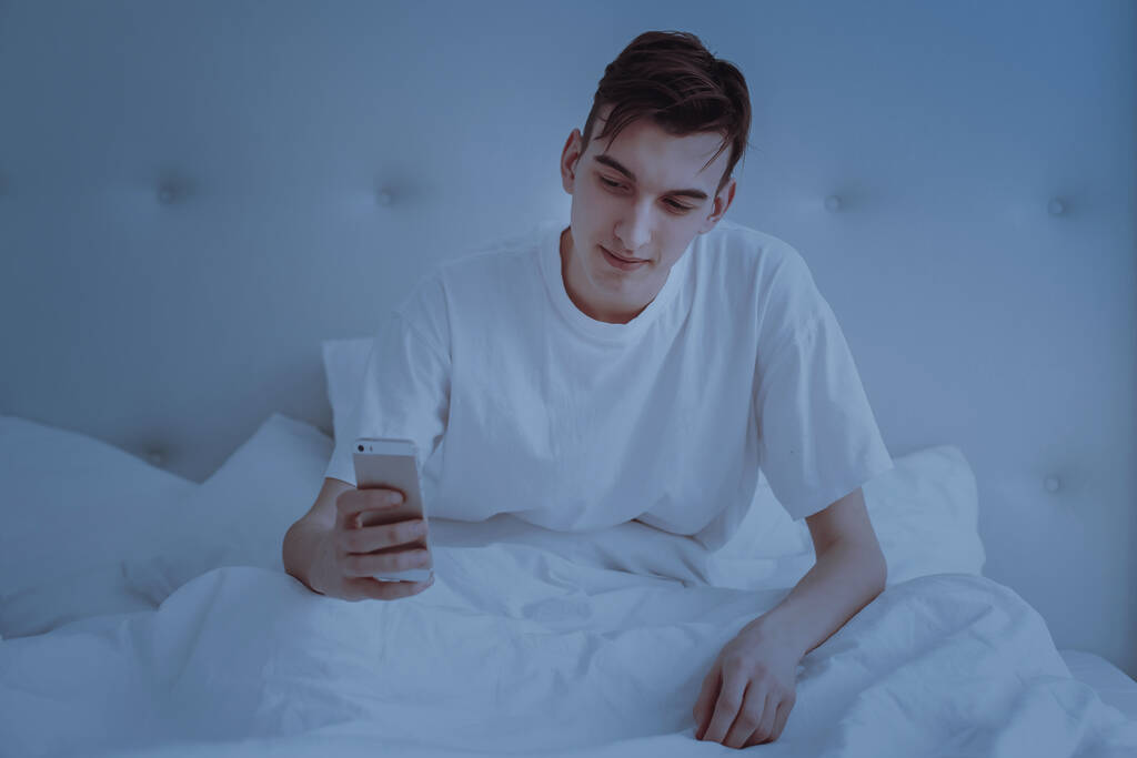 Een knappe jongeman in pyjama die een mobieltje gebruikt terwijl hij 's morgens in bed ligt. Kopieerruimte. - Foto, afbeelding