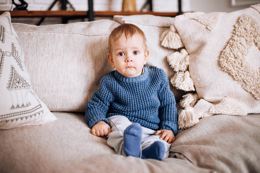 un niño pequeño se sienta en un sofá en un suéter de punto azul
 - Foto, Imagen