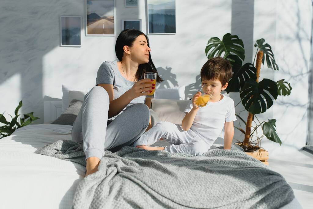 Mooie vrouw met haar zoon die 's morgens sap drinkt in bed. Gezond eten concept - Foto, afbeelding