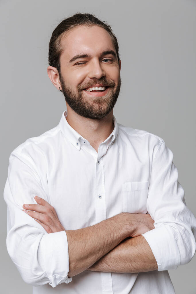 Образ молодого усміхненого оптимістичного красивого бородатого чоловіка, який позує ізольовано на сірому тлі стіни, підморгуючи вам
. - Фото, зображення