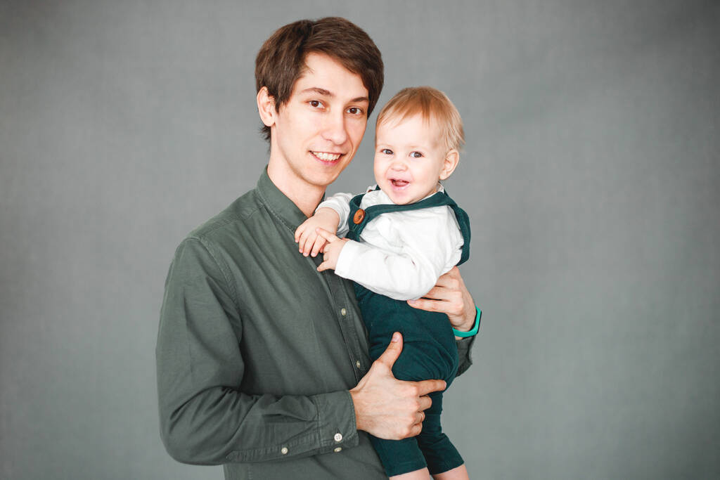 1歳の息子が父親の腕の中に灰色の背景で座り - 写真・画像
