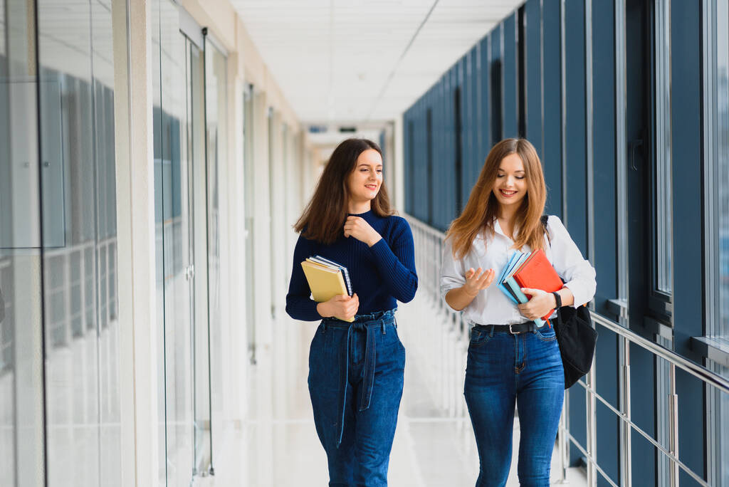 Koridorda kitap ve çantalarla dikilen iki genç kız öğrenci birbirleriyle konuşuyor.. - Fotoğraf, Görsel