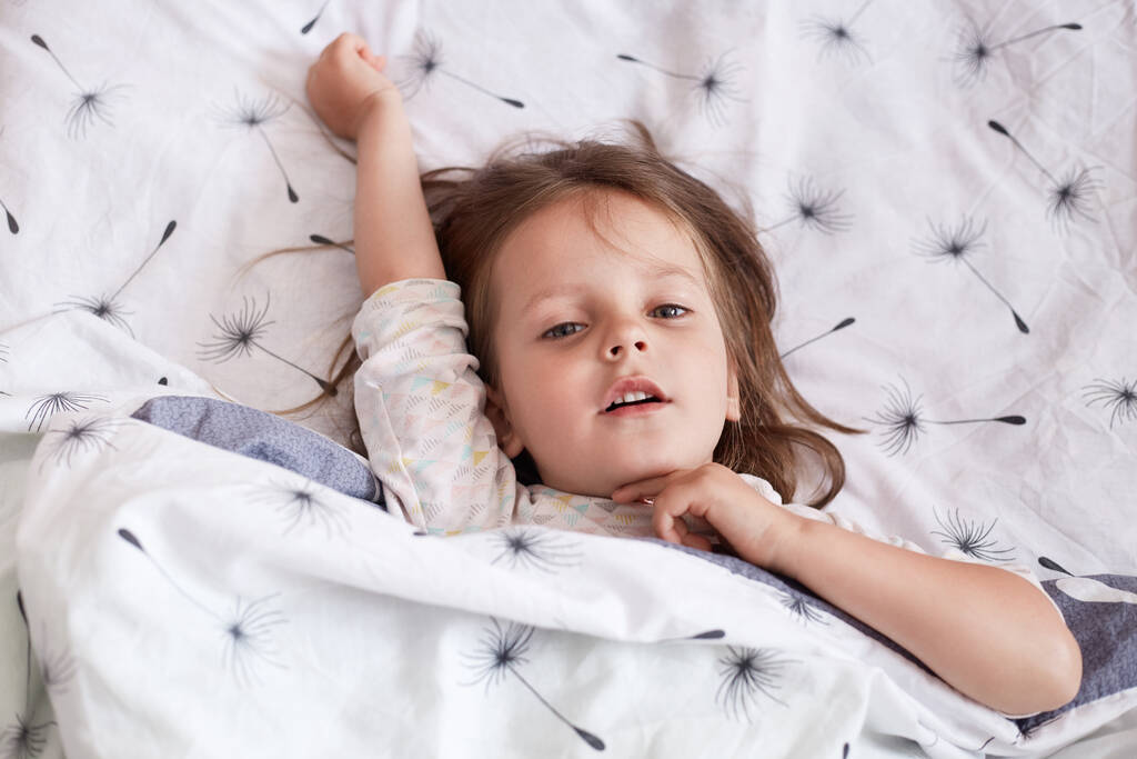 Горизонтальний внутрішній портрет прекрасної чарівної маленької дитини, що піднімає одну руку, має мирне вираження обличчя, лежить у ліжку, насолоджується вільним часом, дивлячись прямо на камеру. Концепція дитинства
. - Фото, зображення