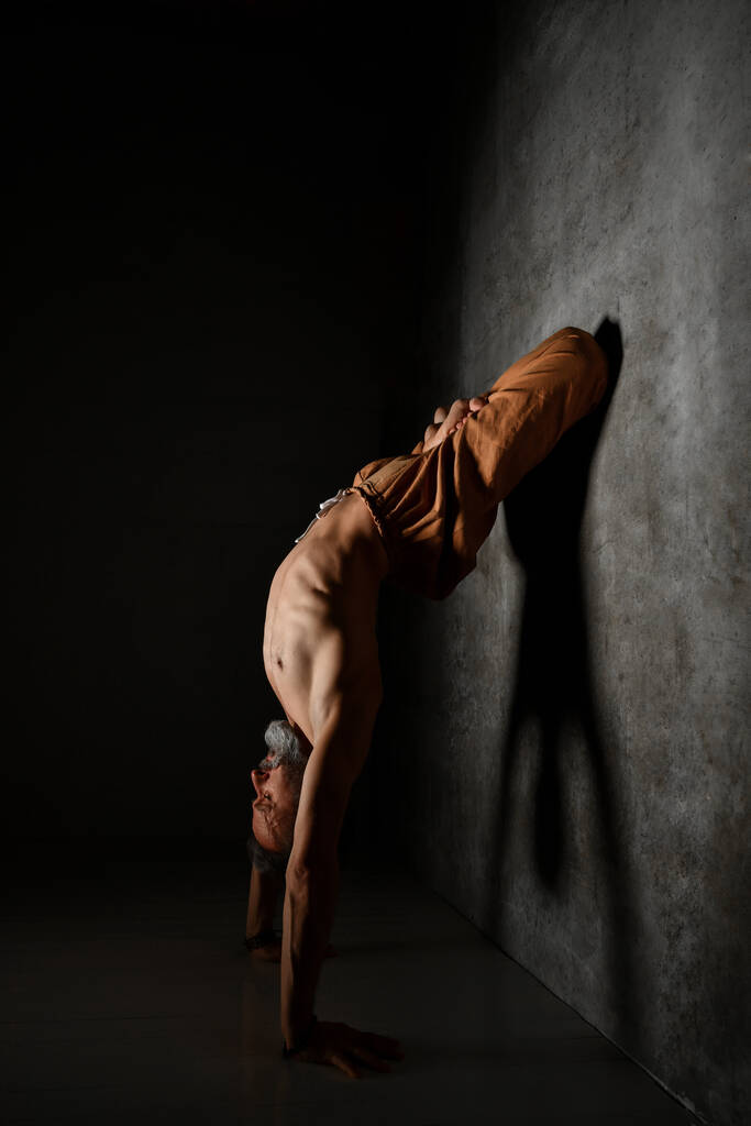 Maschio anziano con torso nudo, in pantaloni marroni sta praticando yoga. In piedi sulle mani, appoggiata alla parete da gambe in posa di loto - Foto, immagini