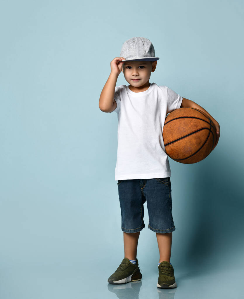 白いTシャツ、グレーキャップ、デニムショートパンツ、カーキスニーカーの小さな子供。バスケットボールボールを持ち笑顔で青い背景にポーズをとって - 写真・画像