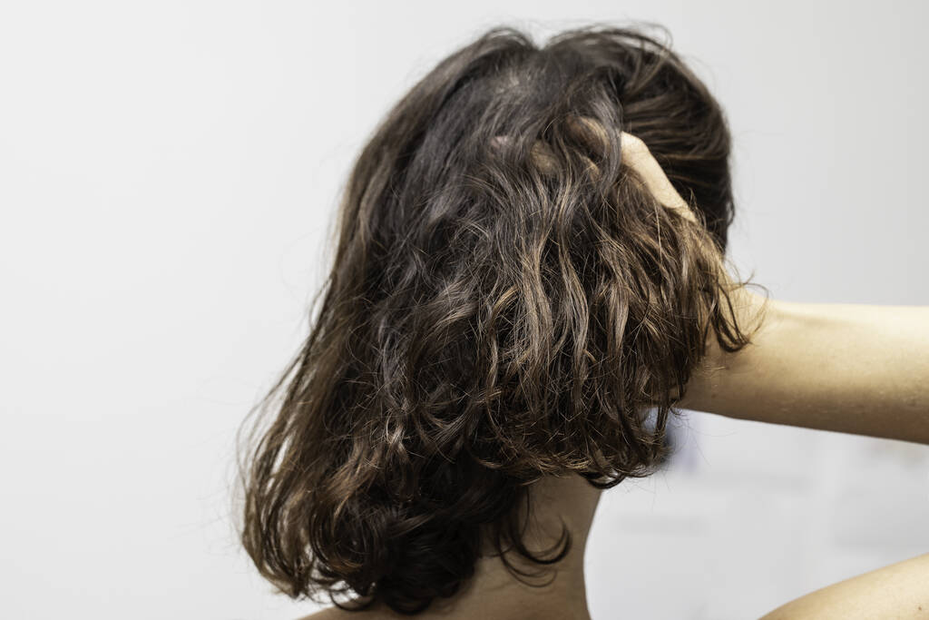 Una bella donna bruna con capelli ondulati di lunghezza della spalla è vista da vicino dalla schiena, isolata su uno sfondo bianco mentre passa le dita tra i capelli con spazio per copiare
. - Foto, immagini