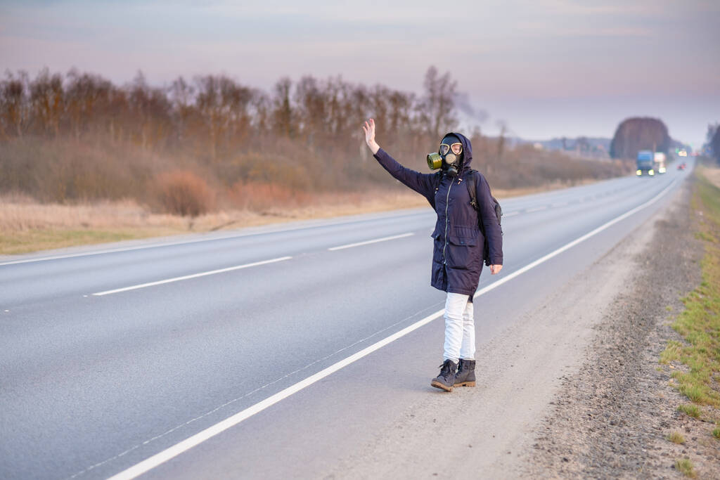 COVID-19. Une fille en tenue sombre, un masque à gaz et une cagoule sur la tête. Journée nuageuse à la campagne. Elle lève la main et tente d'arrêter la voiture qui passe. S'échapper d'une zone infectée par un coronavirus
. - Photo, image