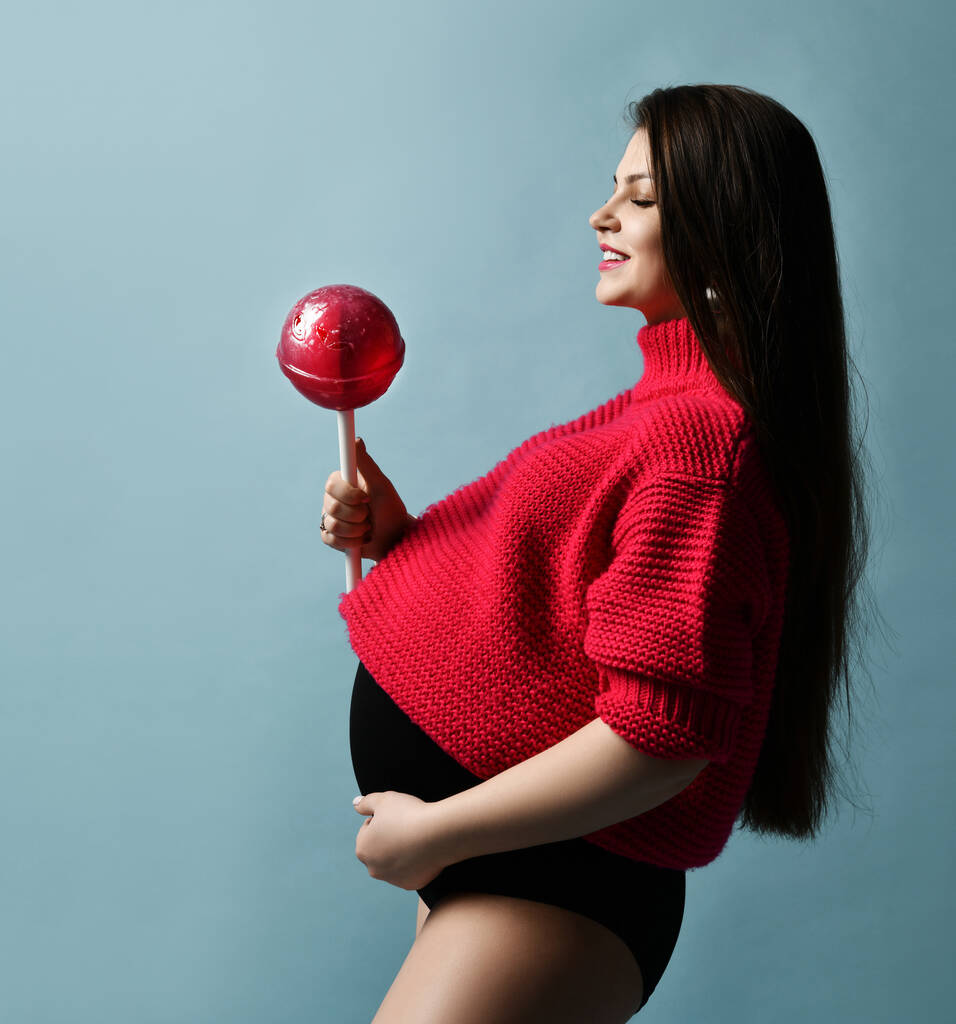 Gyönyörű terhes lány piros pulóverben egy hatalmas nyalókát tart pasztell kéken pózolva. - Fotó, kép