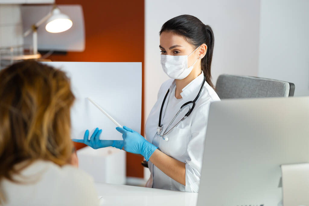 Σοβαρή γυναίκα γιατρός με ιατρική μάσκα που δείχνει κάτι σε χαρτί στο νοσοκομείο - Φωτογραφία, εικόνα