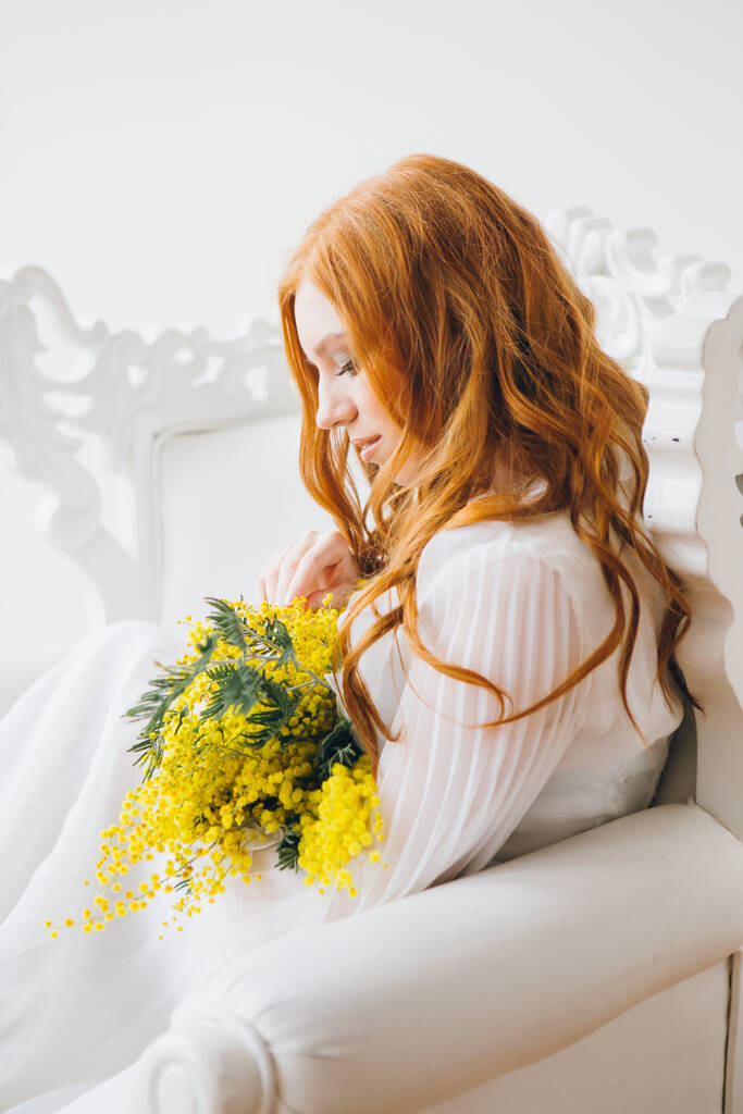 Retrato de uma menina bonita de cabelos vermelhos com uma Mimosa em um vestido branco longo. Concerto da Primavera. Tonificação
. - Foto, Imagem