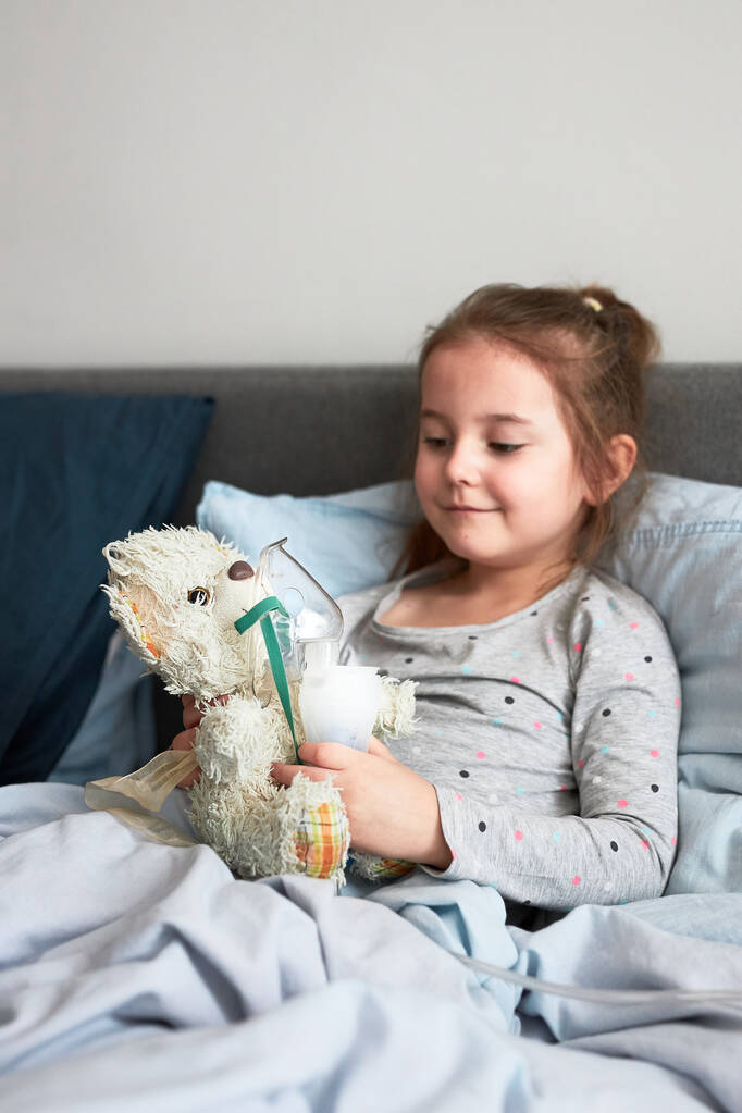 Nemocné dítě se zotavuje v posteli. Dívka hraje tím, že aplikuje lékařské inhalační léčbu s rozprašovačem na svého medvídka - Fotografie, Obrázek