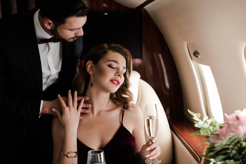 κομψός άντρας αγγίζει σέξι γυναίκα κρατώντας ένα ποτήρι σαμπάνια στο αεροπλάνο - Φωτογραφία, εικόνα