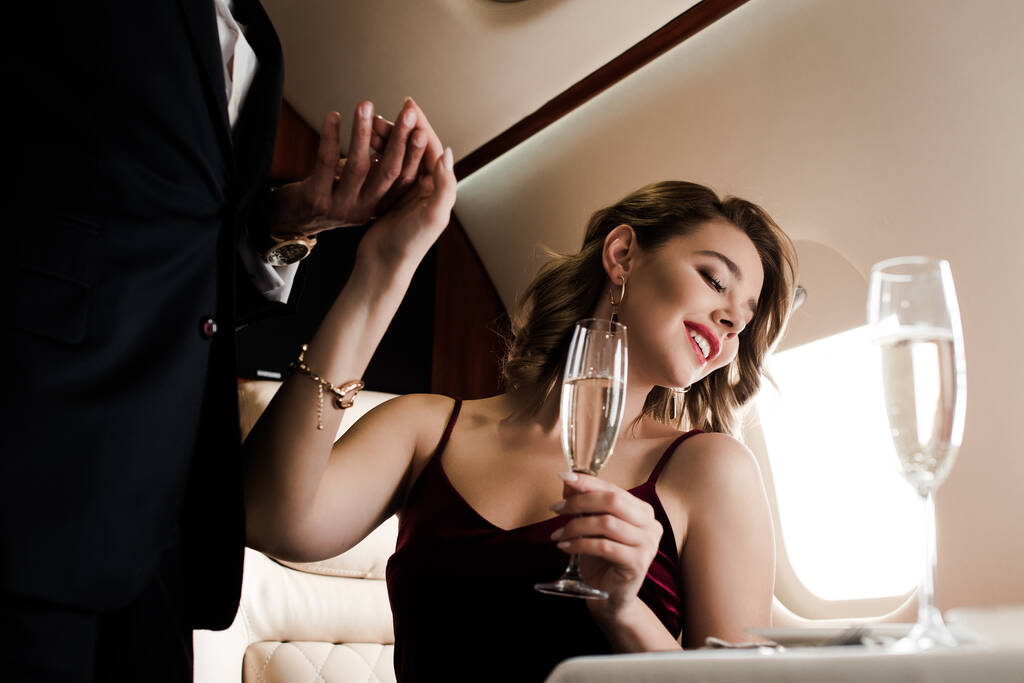 обрізаний вид чоловіка тримає руку усміхненої, привабливої жінки, що тримає келих шампанського в літаку
 - Фото, зображення