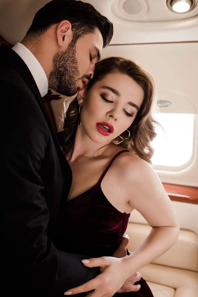 όμορφος άντρας αγκαλιάζει σέξι, κομψή γυναίκα στο αεροπλάνο - Φωτογραφία, εικόνα