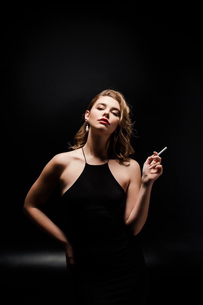 Siyah arka planda poz verirken elinde sigara tutan zarif elbiseli çekici bir kadın. - Fotoğraf, Görsel