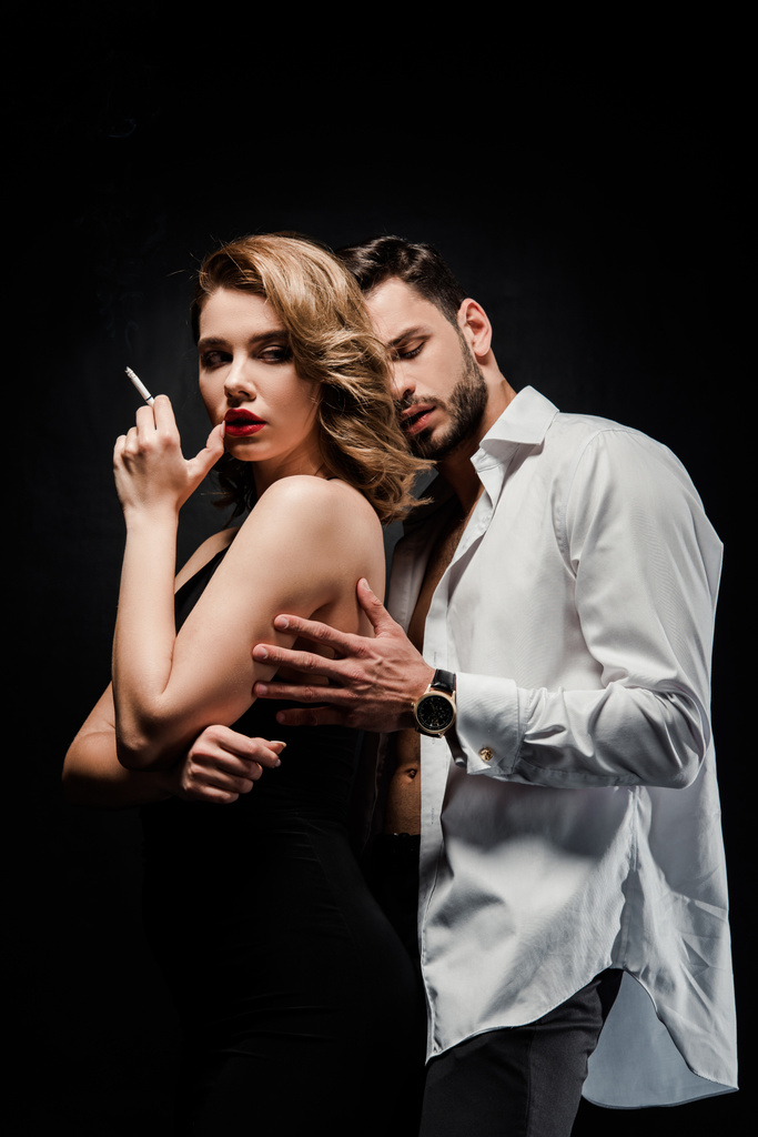 Όμορφος άντρας με λευκό πουκάμισο αγγίζει αυτοπεποίθηση, σαγηνευτική γυναίκα με τσιγάρο απομονωμένο σε μαύρο - Φωτογραφία, εικόνα