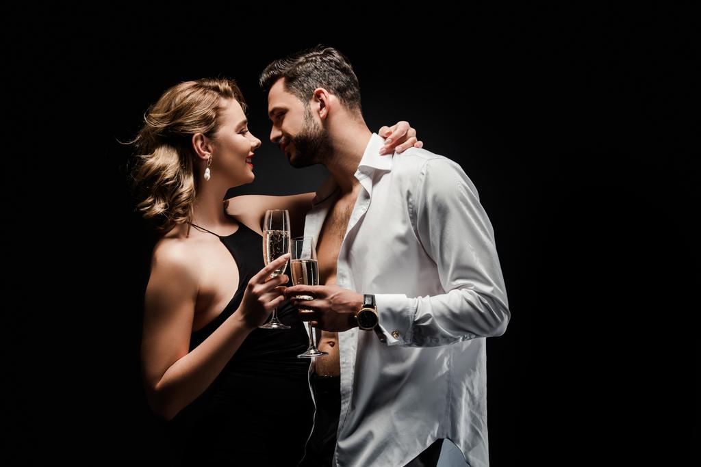 sexy hombre en camisa blanca desabotonada y elegante, seductora mujer sosteniendo copas de champán aisladas en negro
 - Foto, Imagen