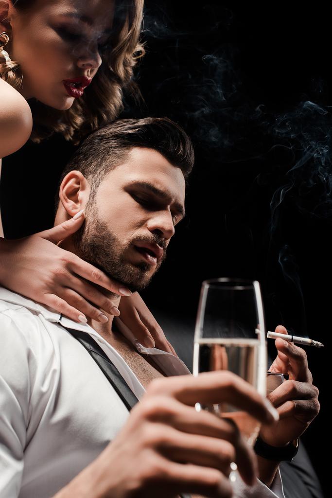 σέξι κορίτσι που αγγίζει το λαιμό του όμορφου άντρα με αυτοπεποίθηση κρατώντας το ποτήρι σαμπάνιας και το κάπνισμα απομονωμένο σε μαύρο - Φωτογραφία, εικόνα