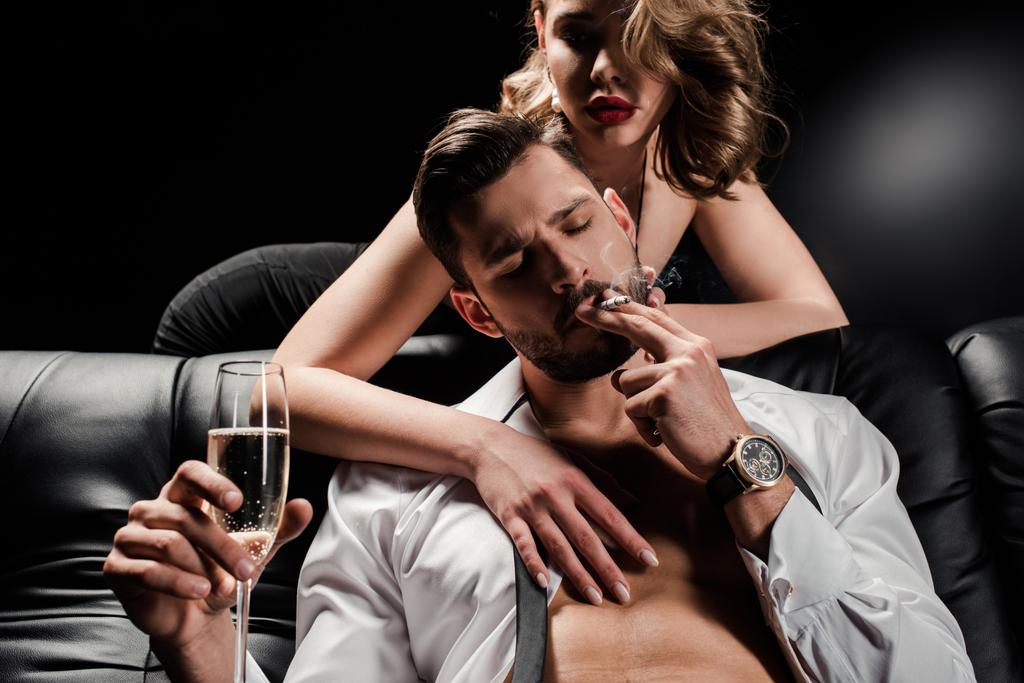 Seksi kadın yakışıklı, kendine güvenen adama dokunuyor. Kanepede oturuyor, elinde şampanya bardağı ve siyah arka planda sigara içiyor. - Fotoğraf, Görsel