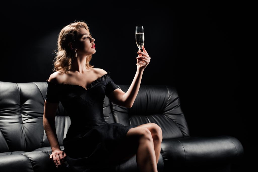 elegáns lány fekete ruhában kezében pohár pezsgő, miközben ül a bőr kanapén elszigetelt fekete - Fotó, kép