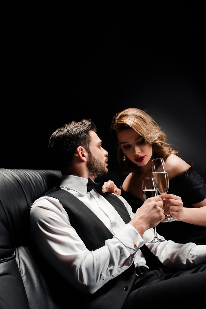 attractive woman and elegant man clinking champagne glasses while sitting on leather sofa on black background - Photo, Image