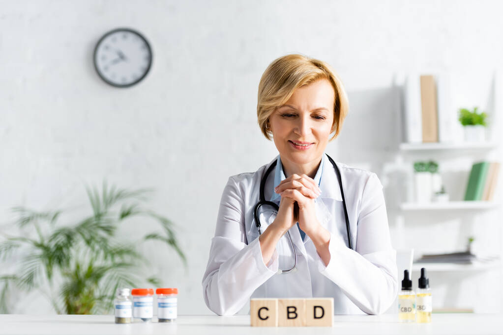 白いコートの幸せな医者は薬で瓶の近くにcbdレタリングとキューブを見て  - 写真・画像