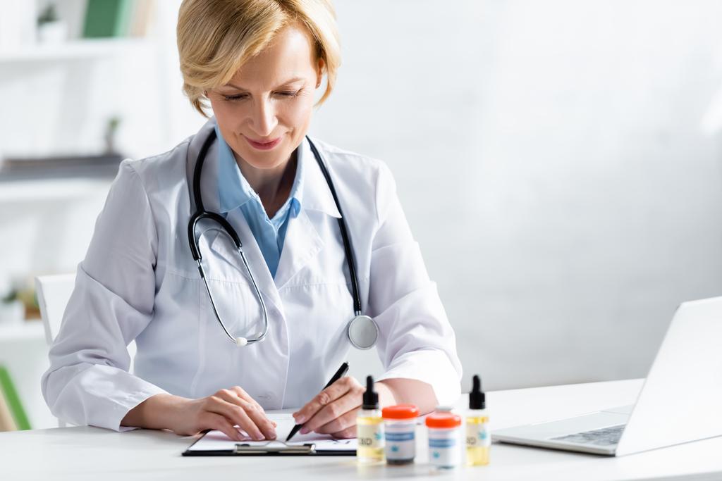 selective focus of cheerful and mature doctor writing prescription near bottles with medical cannabis and cbd  - Photo, Image