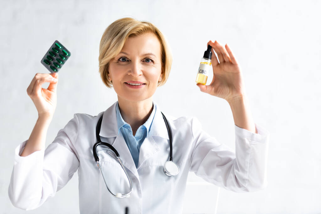 boldog és érett orvos fehér köpenyben tartó palack cbd felirattal és buborékcsomagolás tablettákkal a klinikán  - Fotó, kép