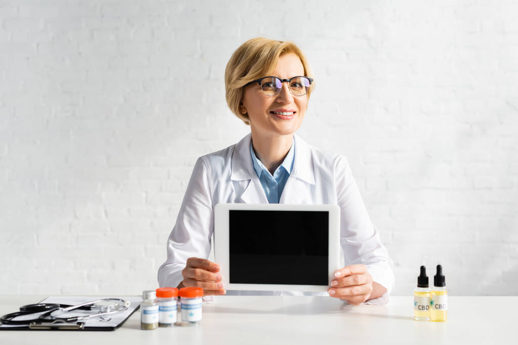gelukkige dokter van middelbare leeftijd met digitale tablet met blanco scherm in de buurt van flessen met cbd en medische cannabis belettering in kliniek  - Foto, afbeelding