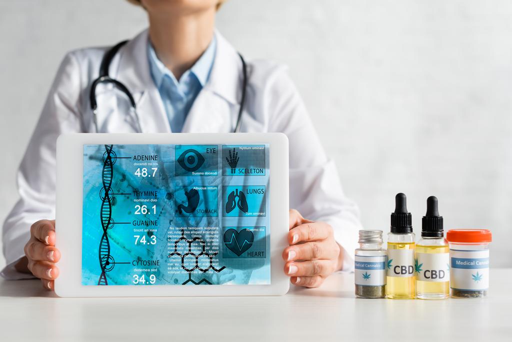 cropped view of mature doctor in white coat holding digital tablet with medical app near bottles with cbd and cannabis lettering  - Photo, Image