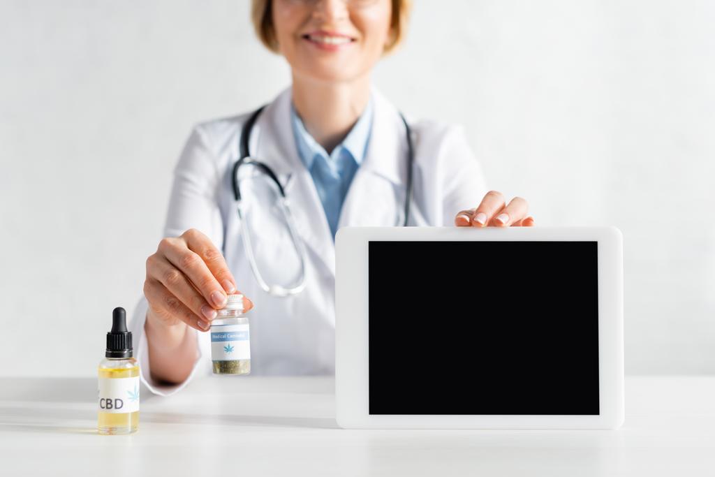 vista cortada de médico feliz e maduro em casaco branco segurando tablet digital com tela em branco e garrafa com letras cannabis medicinal
  - Foto, Imagem