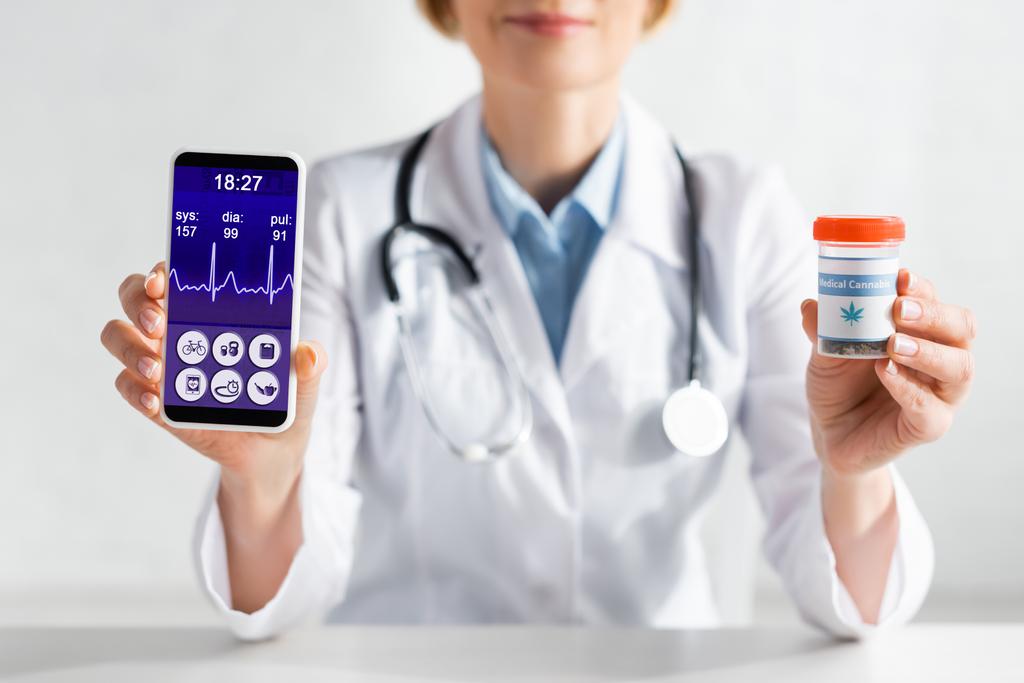 bijgesneden weergave van volwassen arts in witte jas met smartphone met hartslag en fles met medische cannabis belettering  - Foto, afbeelding