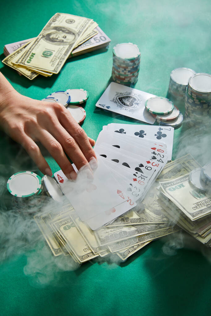 Обрезанный вид женской руки с игральными картами, деньгами и жетонами казино с дымом вокруг на зеленый
 - Фото, изображение