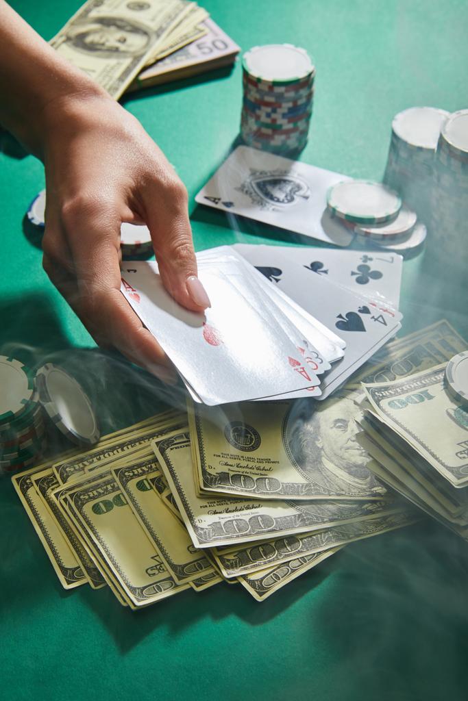 Cropped view of woman holding playing cards near dollar banknotes and casino tokens with smoke around on green  - Photo, Image