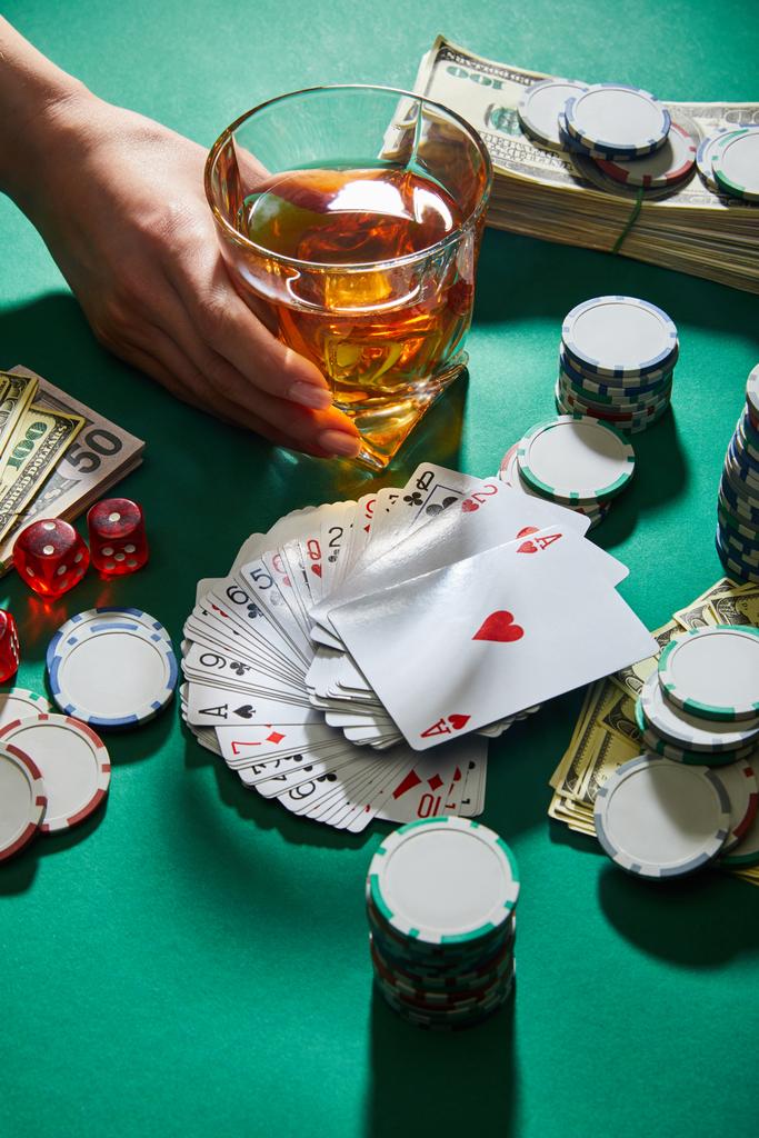 Ausgeschnittene Ansicht einer Frau mit einem Glas Cognac in der Nähe von Geld, Spielkarten, Würfeln und Casino-Wertmarken auf grün - Foto, Bild