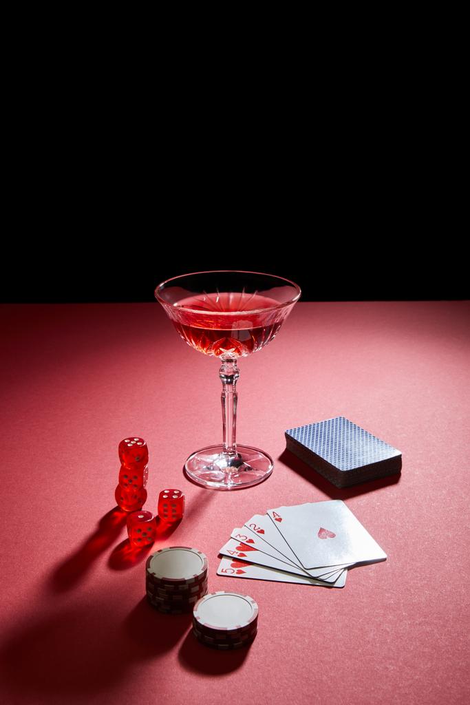 Ποτήρι κοκτέιλ κοντά σε κάρτες παιχνιδιού, μάρκες καζίνο και ζάρια σε κόκκινη επιφάνεια που απομονώνονται σε μαύρο - Φωτογραφία, εικόνα