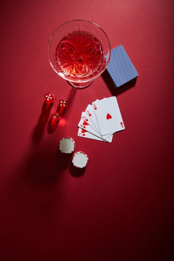 Oyun kartları, kumarhane fişleri ve kırmızı arka plandaki zar yakınlarındaki kokteyl kadehinin üst görüntüsü - Fotoğraf, Görsel