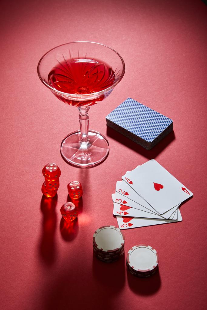 Vista de alto ángulo del vaso de cóctel cerca de jugar a las cartas, fichas de casino y dados en el fondo rojo
 - Foto, Imagen
