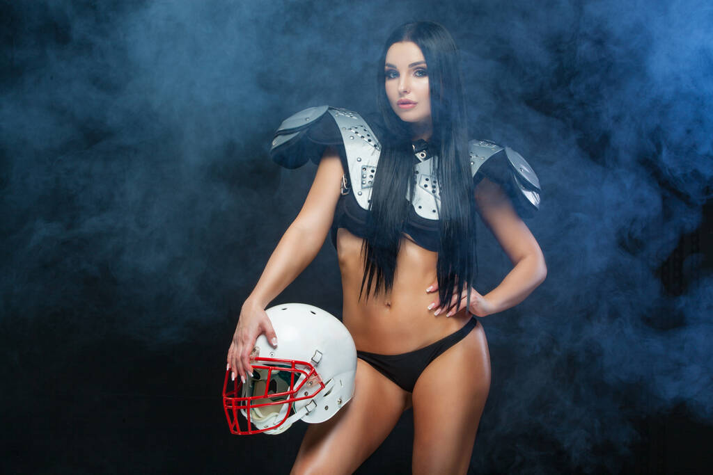 Amerikai foci. Front view fiatal sportos barna visel szexi egyenruhát rögbi labdarúgó pózol sisakkal füst elszigetelt fekete háttér - Fotó, kép