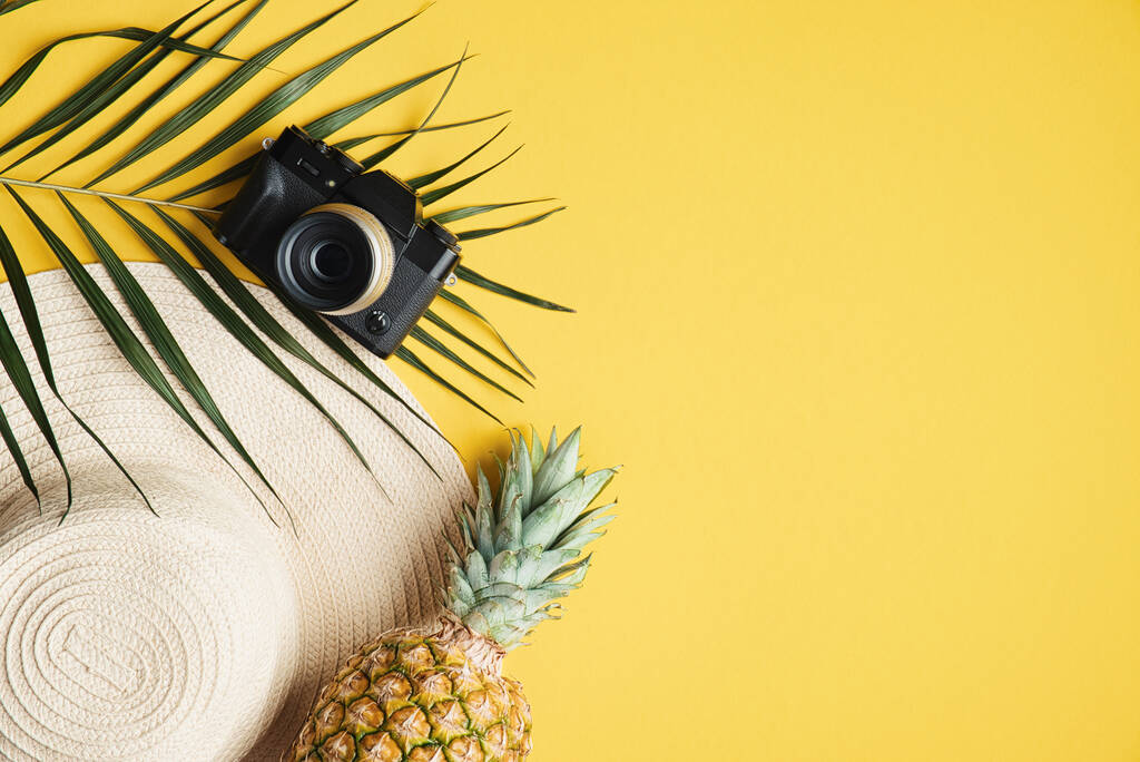 Płaskie leżaki akcesoria podróżnika na żółtym tle ze słomkowym kapeluszem plażowym, ananasem, tropikalnym liściem palmy i aparatem fotograficznym. Top widok wakacje, podróże, koncepcja wakacje - Zdjęcie, obraz