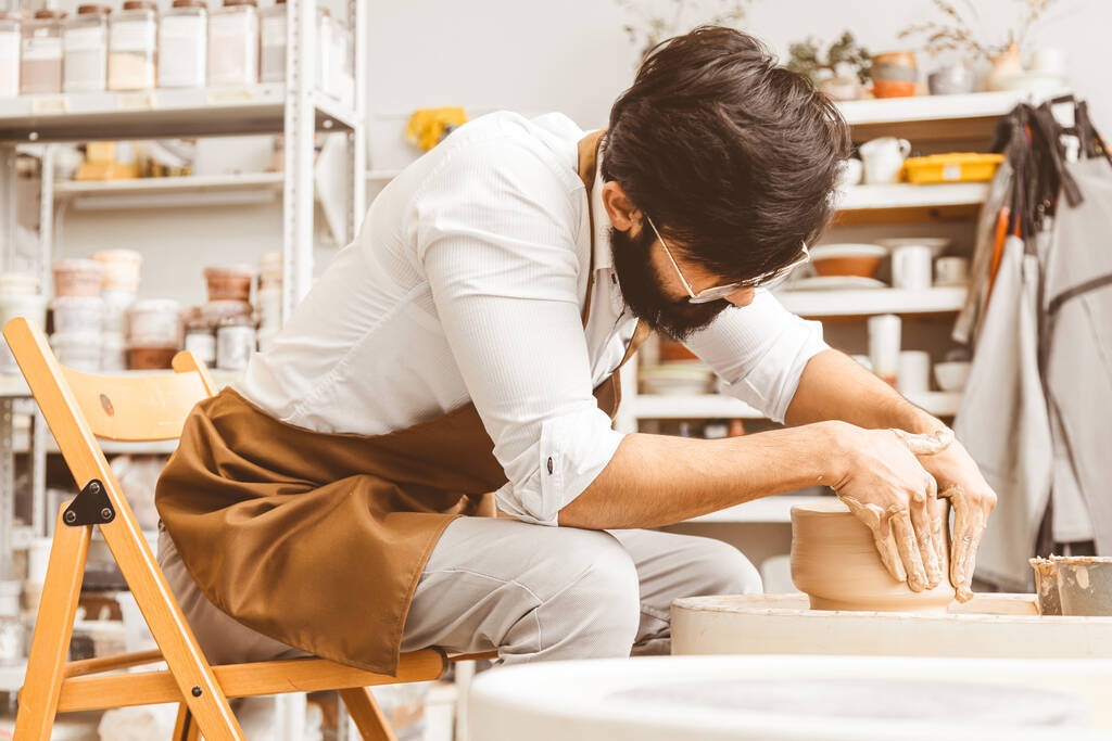 Ein junger männlicher Töpfer bastelt in seiner Werkstatt an einer Töpferscheibe und stellt ein Tonprodukt her - Foto, Bild