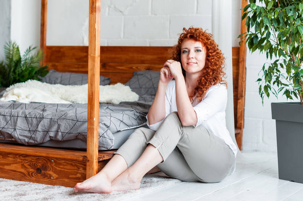 Atraente menina encaracolado ruivo sentado perto de uma cama de madeira
 - Foto, Imagem