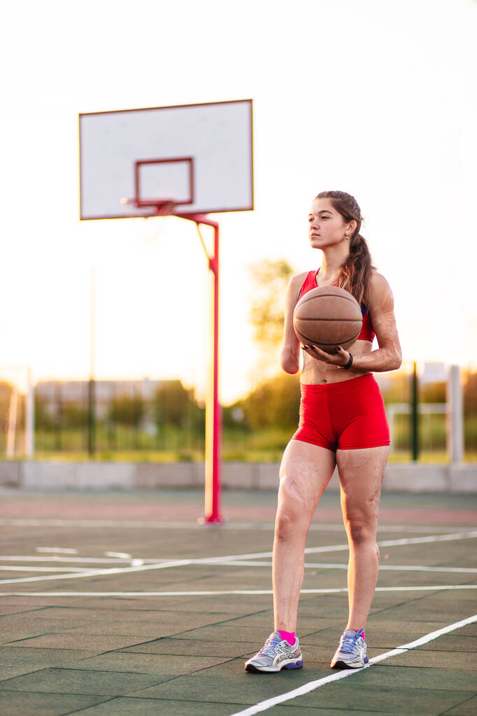 Молода жінка спортсменка з ампутованою рукою і опіками на тілі. Вона тримає в руках баскетбольний м'яч після тренування на відкритому повітрі під час заходу сонця
. - Фото, зображення