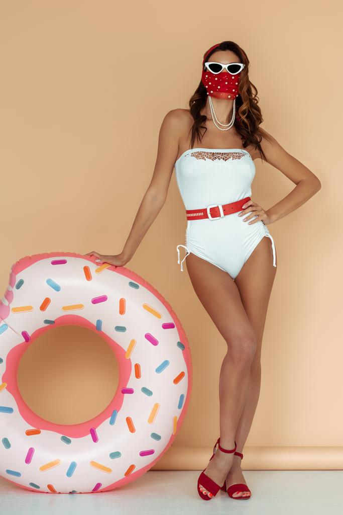 stylish girl in sunglasses, mask and bathing suit standing with hand on hip near inflatable ring on beige - Photo, Image