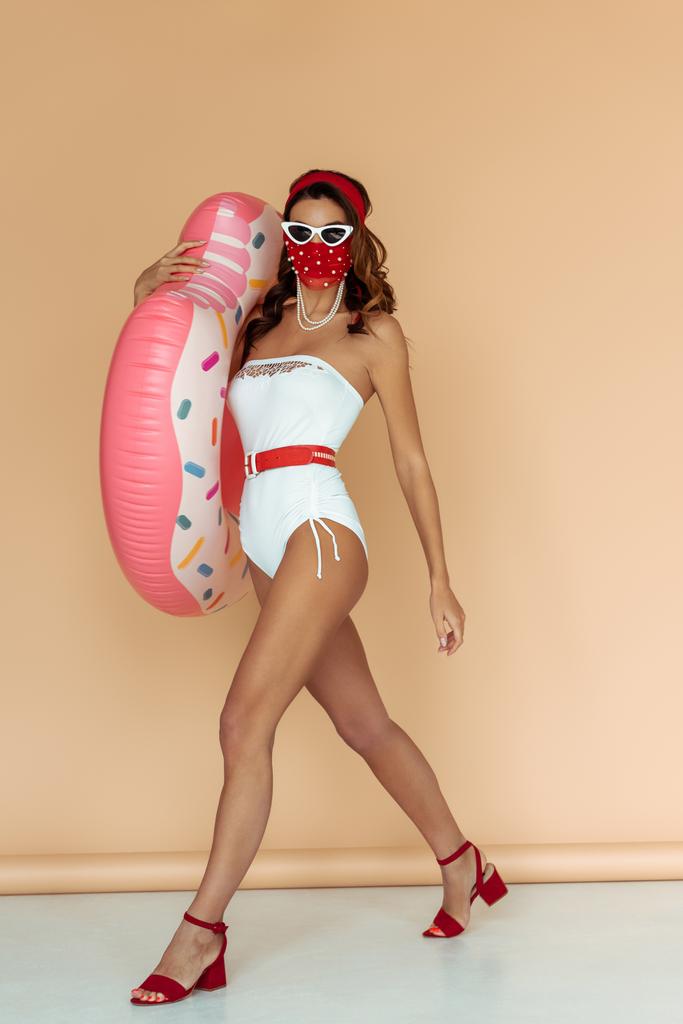 stylish girl in sunglasses, mask and bathing suit holding inflatable ring while walking on beige - Photo, Image