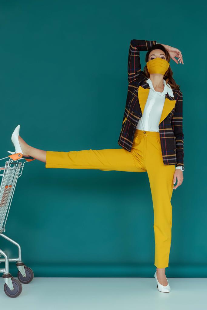 donna alla moda in maschera gialla mettendo gamba sul carrello della spesa vuoto su blu
  - Foto, immagini