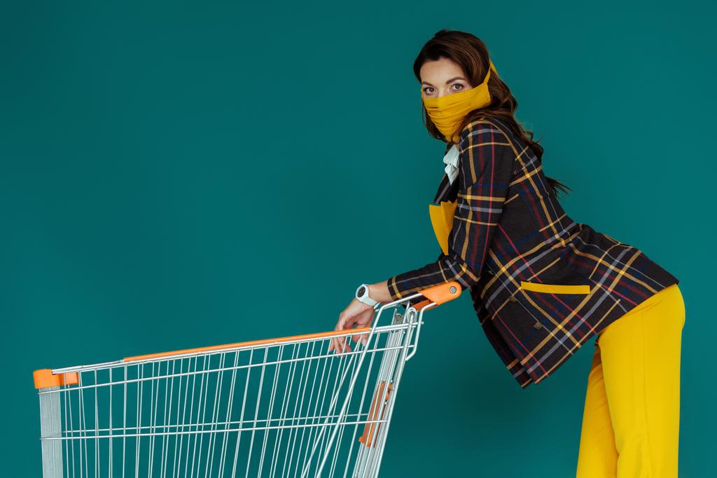 mujer con estilo en máscara amarilla de pie con el carrito de compras vacío en azul
 - Foto, Imagen