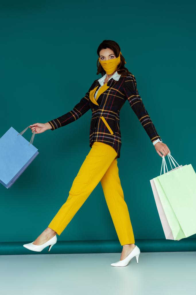 κομψή γυναίκα με κίτρινη μάσκα και καρό σακάκι περπάτημα με τσάντες για ψώνια σε μπλε - Φωτογραφία, εικόνα