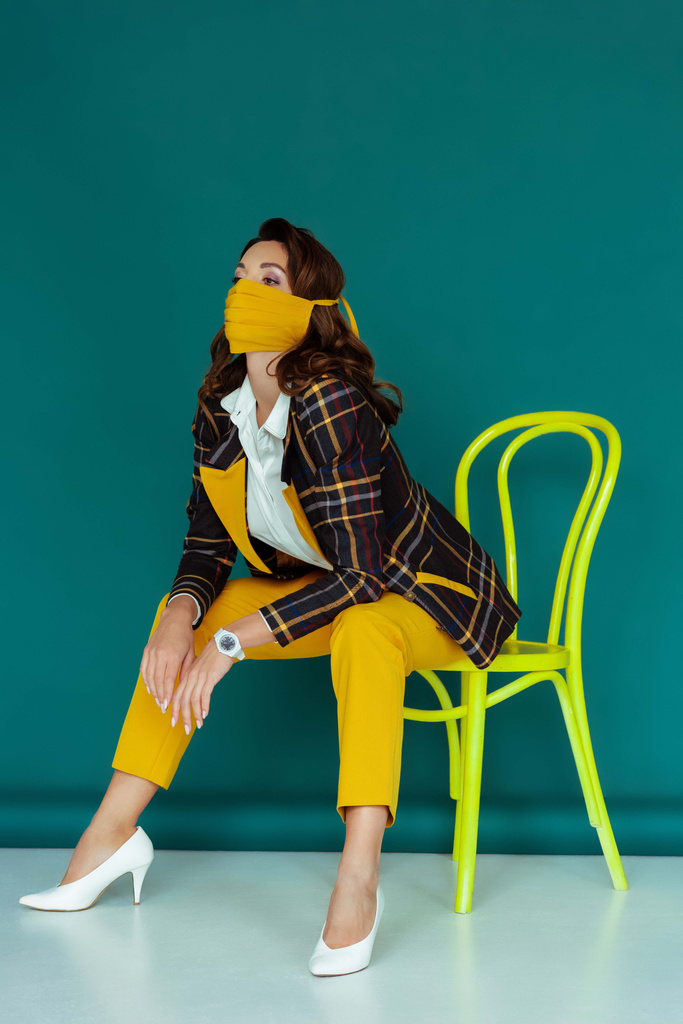 trendy woman in yellow mask and plaid blazer posing while sitting on chair on blue - Photo, Image